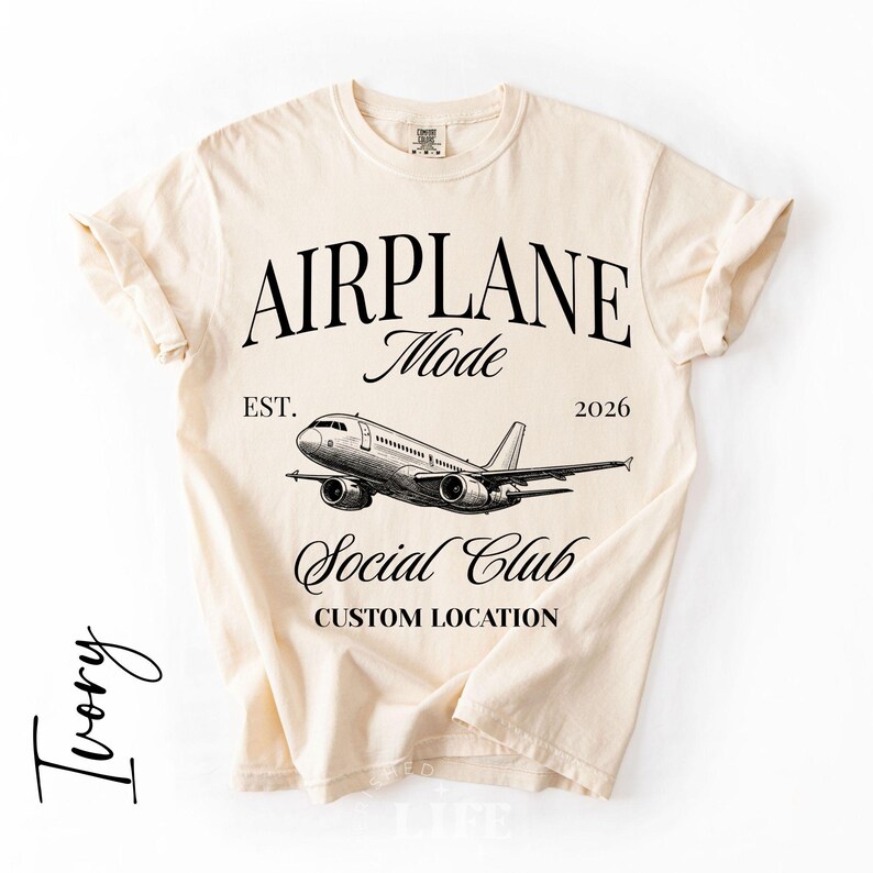Puede incluir: Camiseta de color marfil con las palabras "AIRPLANE Mode Social Club" y una ilustraci&oacute;n detallada de un avi&oacute;n. La camiseta tambi&eacute;n incluye el texto "EST. 2026" y "CUSTOM LOCATION".