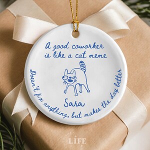 May include: A white ceramic ornament with a gold string for hanging. The ornament features blue text and a cat illustration. The text reads: "A good coworker is like a cat meme. Doesn't fix anything, but makes the day better. Sara."