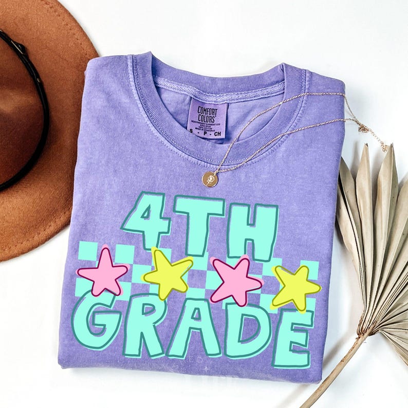 May include: A lavender Comfort Colors t-shirt with the words "4TH GRADE" in turquoise, outlined in white, and a checkerboard pattern. Pink and yellow stars are above the word "GRADE". A gold necklace is draped over the shirt.