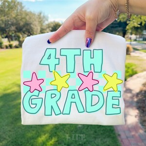 May include: A white t-shirt with the words "4TH GRADE" in a teal font, outlined in white. Pink and yellow stars are above the word "GRADE". The shirt is held up by a hand with blue and purple nail polish.