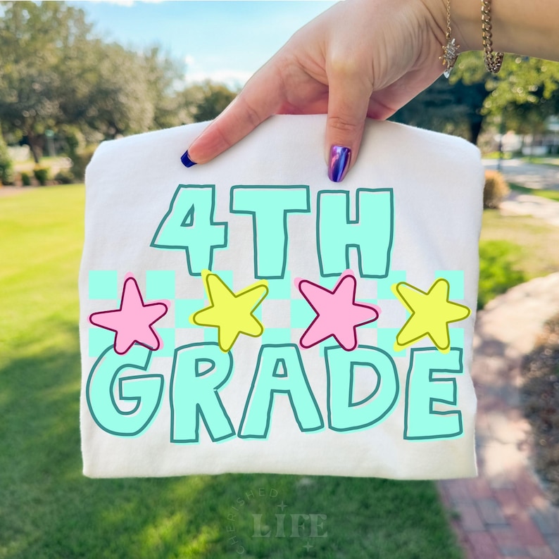 May include: A white t-shirt with the words "4TH GRADE" in a teal font, outlined in white. Pink and yellow stars are above the word "GRADE". The shirt is held up by a hand with blue and purple nail polish.