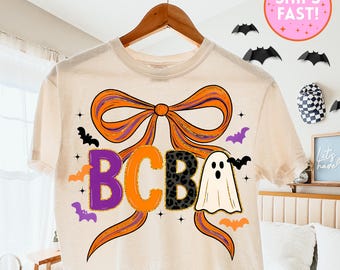 May include: Cream-colored t-shirt with a large orange and purple bow design. The shirt features the letters "BCBA" in purple, orange, leopard print, and a ghost graphic. Bats and the text "SHIPS FAST!" are also visible.