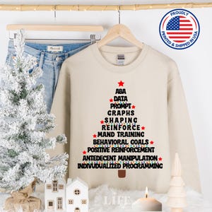 May include: A beige sweatshirt with a Christmas tree design featuring the words "ABA, Data Prompt, Graphs, Shaping, Reinforce, Mand Training, Behavioral Goals, Positive Reinforcement, Antecedent Manipulation, Individualized Programming". The tree is decorated with colorful lights.