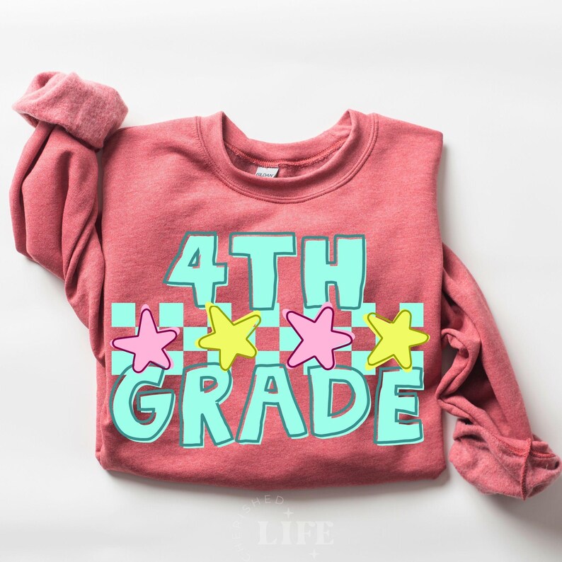 May include: A red-orange sweatshirt with the words "4TH GRADE" in a turquoise font with a white checkered background. Pink and yellow star graphics are above the word "GRADE". The sweatshirt has long sleeves.