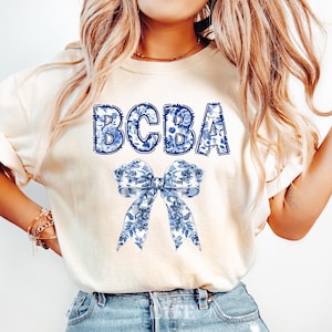 May include: Cream-colored t-shirt featuring the letters "BCBA" in a blue floral design. Below the text is a large bow with a matching blue floral pattern. The shirt is a casual style.