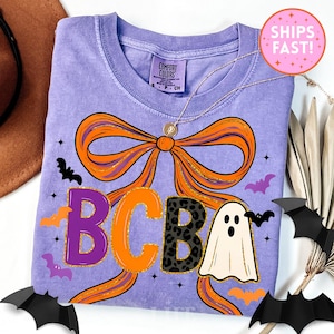 May include: A lilac Comfort Colors t-shirt featuring a large orange bow with gold glitter accents. The shirt has the letters "BCBA" in purple, orange, and leopard print, along with a ghost and bats, perfect for Halloween.