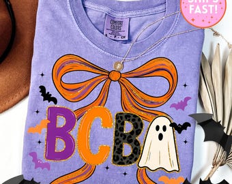 May include: A lilac Comfort Colors t-shirt featuring a large orange bow with gold glitter accents. The shirt has the letters "BCBA" in purple, orange, and leopard print, along with a ghost and bats, perfect for Halloween.