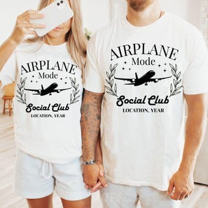 May include: Two white t-shirts with the text "AIRPLANE Mode Social Club LOCATION, YEAR" in black. Each shirt features a silhouette of an airplane inside a laurel wreath. The shirts are worn by two people.