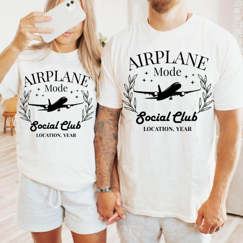 May include: Two white t-shirts with the text "AIRPLANE Mode Social Club LOCATION, YEAR" in black. Each shirt features a silhouette of an airplane inside a laurel wreath. The shirts are worn by two people.