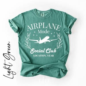 May include: A light green t-shirt with the words "AIRPLANE Mode Social Club" in white, along with an airplane graphic. The shirt has rolled-up sleeves and a relaxed fit. The text "Light Green" is written in cursive on the left side.