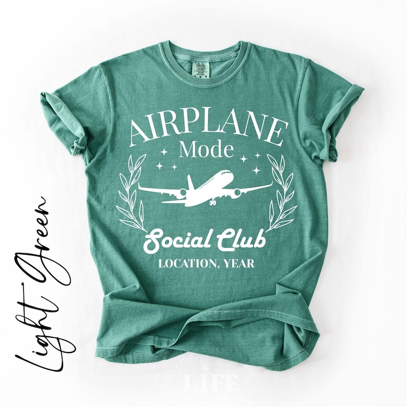 May include: A light green t-shirt with the words "AIRPLANE Mode Social Club" in white, along with an airplane graphic. The shirt has rolled-up sleeves and a relaxed fit. The text "Light Green" is written in cursive on the left side.