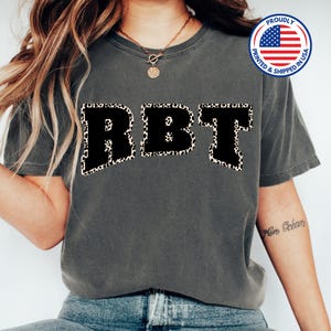May include: A gray t-shirt with the letters "RBT" in black with a leopard print border. The shirt is being worn by a person. The text "Proudly Printed & Shipped in USA" is on a small American flag sticker in the top right corner of the image.