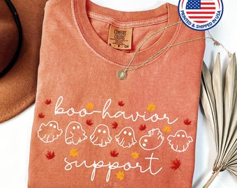 May include: A burnt orange t-shirt with a white graphic that says "boo-havior support" with a row of ghosts and fall leaves. The shirt is made by Comfort Colors and is printed and shipped in the USA.