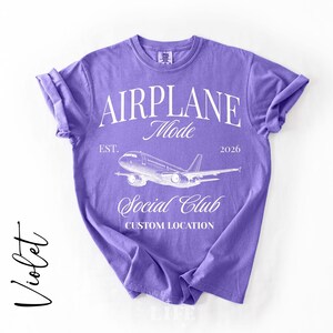 Puede incluir: Camiseta violeta con mangas enrolladas. La camiseta presenta el texto "AIRPLANE Mode Social Club" con un gr&aacute;fico de avi&oacute;n. Tambi&eacute;n se incluye el texto "EST. 2026". La palabra "Violet" est&aacute; escrita en cursiva a la izquierda.