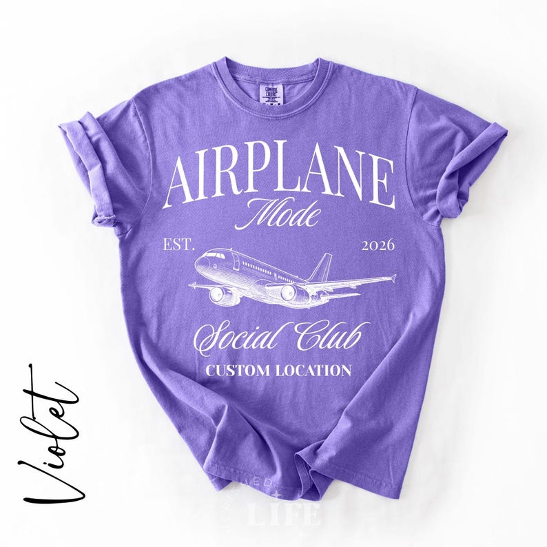 Puede incluir: Camiseta violeta con mangas enrolladas. La camiseta presenta el texto "AIRPLANE Mode Social Club" con un gr&aacute;fico de avi&oacute;n. Tambi&eacute;n se incluye el texto "EST. 2026". La palabra "Violet" est&aacute; escrita en cursiva a la izquierda.