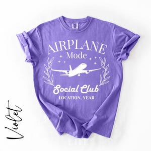 May include: Violet-colored t-shirt with the words "AIRPLANE MODE Social Club" in white, along with an airplane graphic. The shirt has rolled-up sleeves and a crew neck. The word "Violet" is written in cursive on the left side.