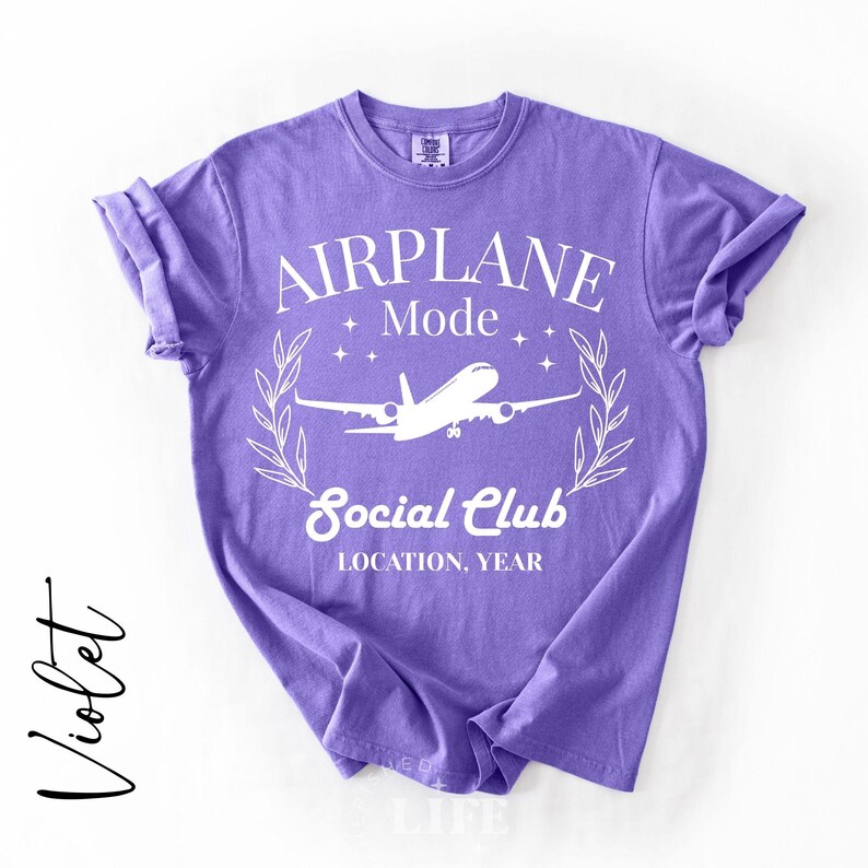 May include: Violet-colored t-shirt with the words "AIRPLANE MODE Social Club" in white, along with an airplane graphic. The shirt has rolled-up sleeves and a crew neck. The word "Violet" is written in cursive on the left side.