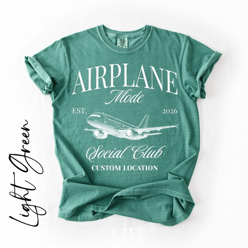 Puede incluir: Camiseta verde claro con las palabras "AIRPLANE Mode" y un gr&aacute;fico de un avi&oacute;n. La camiseta tambi&eacute;n tiene el texto "EST. 2026" y "Social Club". La parte inferior de la camiseta dice "Custom Location".