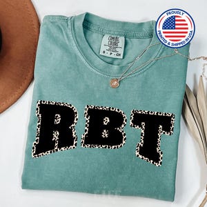 May include: A teal blue t-shirt with the letters RBT in black with a leopard print outline. The shirt is made of a soft, comfortable material.