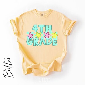 May include: A light yellow t-shirt with the words "4TH GRADE" in turquoise block letters, accented with pink and yellow stars. The shirt is made of a soft material and is likely for children.