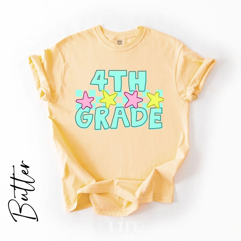 May include: A light yellow t-shirt with the words "4TH GRADE" in turquoise block letters, accented with pink and yellow stars. The shirt is made of a soft material and is likely for children.