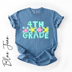 May include: Blue jean t-shirt with the words "4TH GRADE" in a turquoise font with pink and yellow stars. The shirt is a light blue color and has rolled-up sleeves.