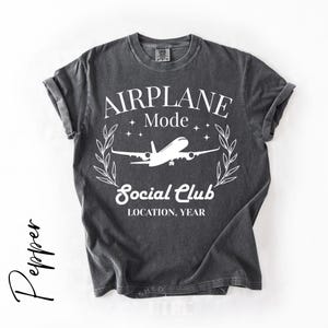 May include: Dark gray t-shirt with rolled-up sleeves. The shirt features a white graphic with the words "AIRPLANE Mode Social Club" and an airplane illustration. The text "LOCATION, YEAR" is also included in the design.