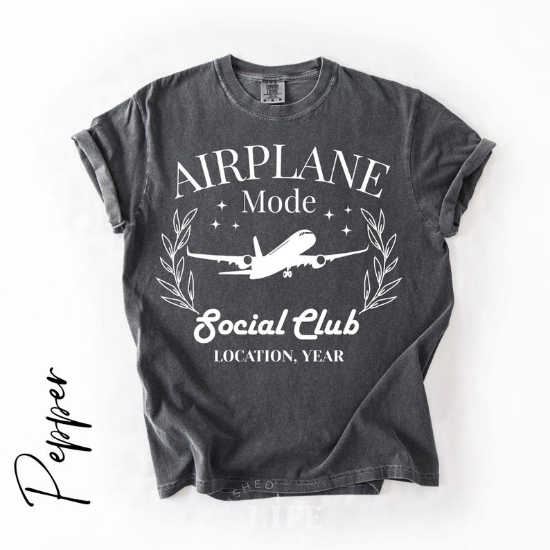 May include: Dark gray t-shirt with rolled-up sleeves. The shirt features a white graphic with the words "AIRPLANE Mode Social Club" and an airplane illustration. The text "LOCATION, YEAR" is also included in the design.