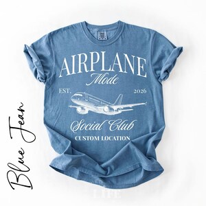 Puede incluir: Camiseta azul vaquera con las palabras "AIRPLANE Mode" y un gr&aacute;fico de avi&oacute;n. La camiseta tambi&eacute;n incluye el texto "Social Club" y "Custom Location". La camiseta es de cuello redondo y tiene mangas enrolladas.