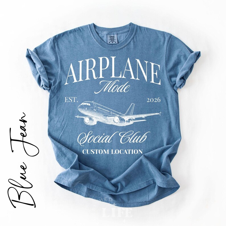 Puede incluir: Camiseta azul vaquera con las palabras "AIRPLANE Mode" y un gr&aacute;fico de avi&oacute;n. La camiseta tambi&eacute;n incluye el texto "Social Club" y "Custom Location". La camiseta es de cuello redondo y tiene mangas enrolladas.