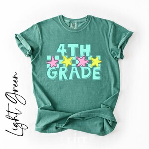 May include: A light green t-shirt with the words "4TH GRADE" in a turquoise font, with pink and yellow stars. The shirt has rolled-up sleeves and a crew neck.
