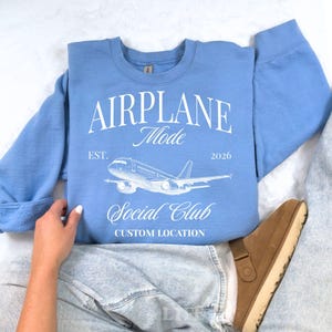 Puede incluir: Sudadera azul claro con las palabras "AIRPLANE Mode Social Club" y un gr&aacute;fico de avi&oacute;n. El texto "EST. 2026" y "CUSTOM LOCATION" tambi&eacute;n est&aacute;n impresos en la sudadera.