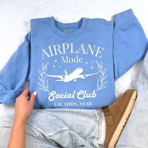 May include: A light blue sweatshirt with the words "AIRPLANE Mode Social Club" in white, along with a graphic of an airplane. The design is encircled by a laurel wreath. The text "LOCATION, YEAR" is also included.