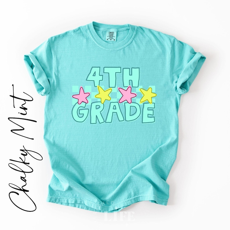 May include: A turquoise t-shirt with rolled-up sleeves. The front features the words "4TH GRADE" in a light green outline, with pink and yellow stars. The words "Chalky Mint" are written in a cursive font on the left side.