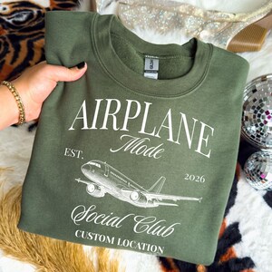 Puede incluir: Sudadera verde oliva con el texto "AIRPLANE Mode Social Club" y un gr&aacute;fico de un avi&oacute;n. El texto tambi&eacute;n incluye "EST. 2026" y "CUSTOM LOCATION" en letras blancas. La sudadera est&aacute; hecha de un material suave.
