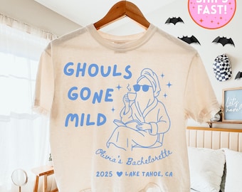 Könnte beinhalten: Cremefarbenes T-Shirt mit blauem Text "GHOULS GONE MILD" und einer Grafik eines Geistes mit Sonnenbrille, Bademantel und Handtuch, der eine Tasse hält. Der Text "Olivia's Bachelorette 2025 Lake Tahoe, CA" ist ebenfalls aufgedruckt.