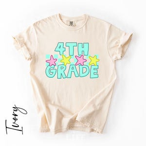 May include: Ivory-colored t-shirt with the words "4TH GRADE" in turquoise, outlined in a darker turquoise. Pink and yellow stars are above the word "GRADE". The shirt has rolled-up sleeves.
