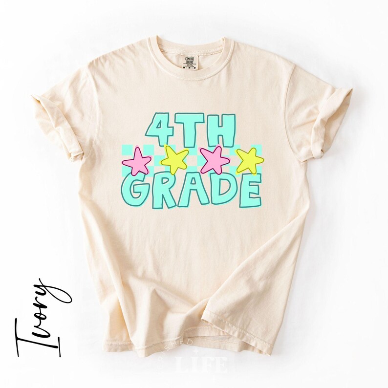 May include: Ivory-colored t-shirt with the words "4TH GRADE" in turquoise, outlined in a darker turquoise. Pink and yellow stars are above the word "GRADE". The shirt has rolled-up sleeves.