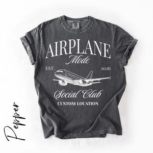 Puede incluir: Camiseta gris oscuro con mangas enrolladas que presenta el texto "AIRPLANE Mode" sobre un gr&aacute;fico de avi&oacute;n. Debajo del gr&aacute;fico, el texto dice "Social Club" y "Custom Location". La camiseta tambi&eacute;n incluye el texto "EST. 2026".
