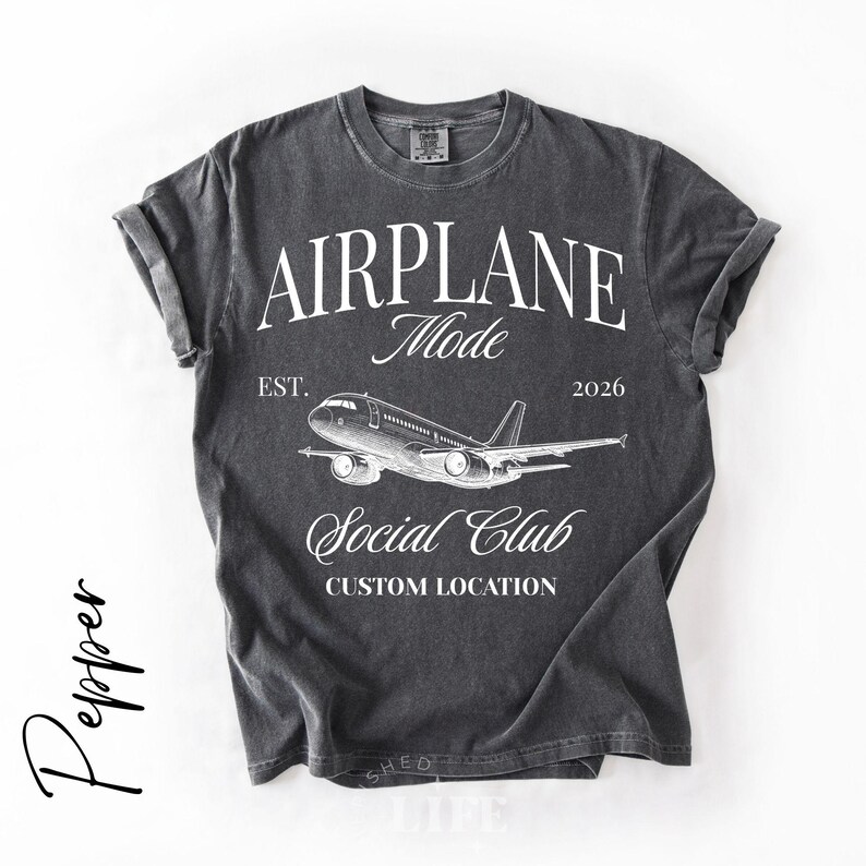 Puede incluir: Camiseta gris oscuro con mangas enrolladas que presenta el texto "AIRPLANE Mode" sobre un gr&aacute;fico de avi&oacute;n. Debajo del gr&aacute;fico, el texto dice "Social Club" y "Custom Location". La camiseta tambi&eacute;n incluye el texto "EST. 2026".
