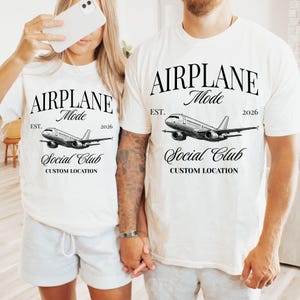 Puede incluir: Dos camisetas blanquecinas con el texto "AIRPLANE Mode" sobre una ilustraci&oacute;n detallada de un avi&oacute;n. Debajo del gr&aacute;fico del avi&oacute;n est&aacute; "Social Club" y "CUSTOM LOCATION". Las camisetas tambi&eacute;n incluyen el texto "EST. 2026".