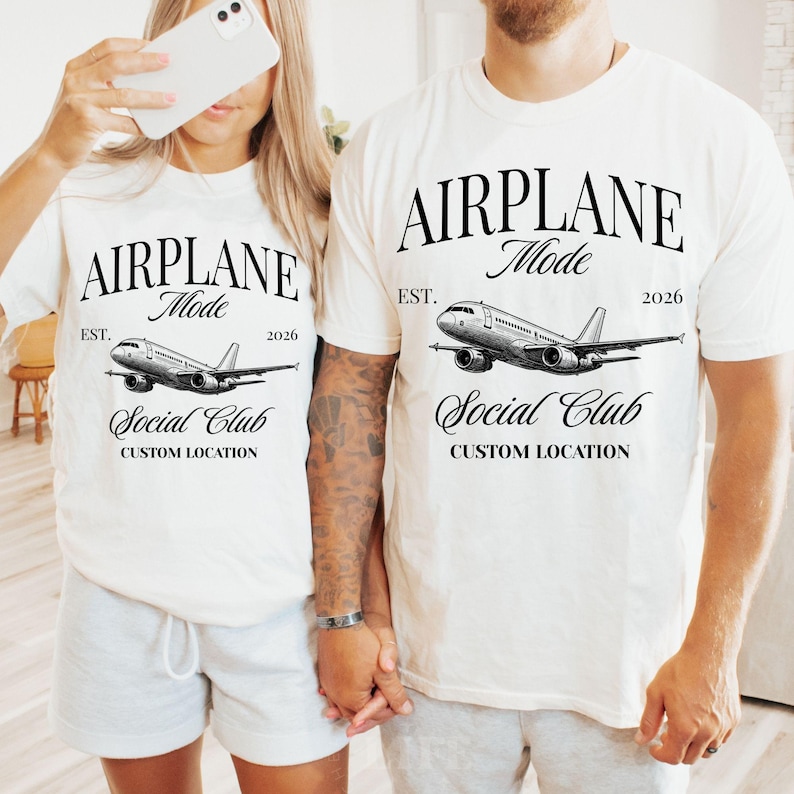 Puede incluir: Dos camisetas blanquecinas con el texto "AIRPLANE Mode" sobre una ilustraci&oacute;n detallada de un avi&oacute;n. Debajo del gr&aacute;fico del avi&oacute;n est&aacute; "Social Club" y "CUSTOM LOCATION". Las camisetas tambi&eacute;n incluyen el texto "EST. 2026".