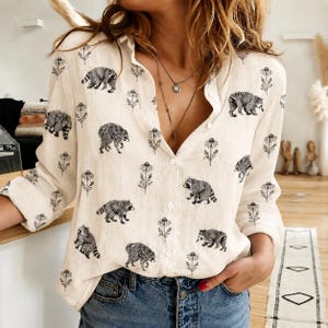May include: A long-sleeved button-down shirt in a light cream color featuring a repeating black and white print of raccoons and tall wildflowers. The shirt has a classic collar, a front button placket, and is paired with blue denim jeans.