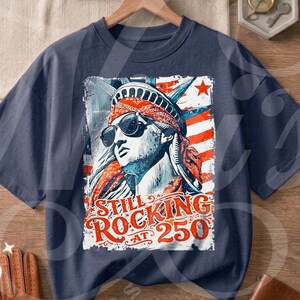 Könnte beinhalten: Marineblaues T-Shirt mit einer Grafik der Freiheitsstatue mit Sonnenbrille und dem Text "Still Rocking at 250." Das Design verwendet rote, weiße und blaue Elemente und erinnert an ein amerikanisches Thema.