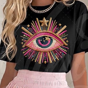 May include: Black t-shirt featuring a colorful embroidered eye design with gold accents and a star. The eye design has pink, blue, and gold details. The model is wearing a pink skirt and gold jewelry. The brand name "Nini's Creations" is visible.