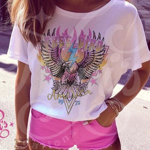 May include: White t-shirt with a graphic of an eagle with the text "Free Bird" and "America 1972" printed on it. The eagle is surrounded by flames and stars.