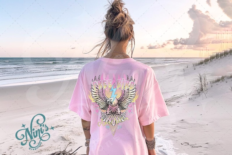 May include: A pink t-shirt with a graphic of a colorful eagle with wings spread and the text "FREE BIRD" on the back. The shirt is being worn by a person on a beach.
