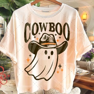 Puede incluir: Camiseta color crema con un dise&ntilde;o de un fantasma con sombrero de vaquero. La palabra "COWBOO" est&aacute; impresa encima del fantasma en una fuente desgastada. Estrellas y detalles en naranja rodean al fantasma y el texto.