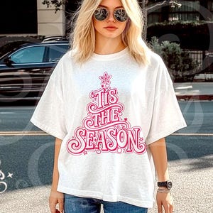 Op de afbeelding: Een wit T-shirt met een roze kerstboomontwerp en de tekst "Tis The Season". Het T-shirt wordt gedragen door een persoon met een zonnebril en een spijkerbroek. Een casual, feestelijk kledingstuk.