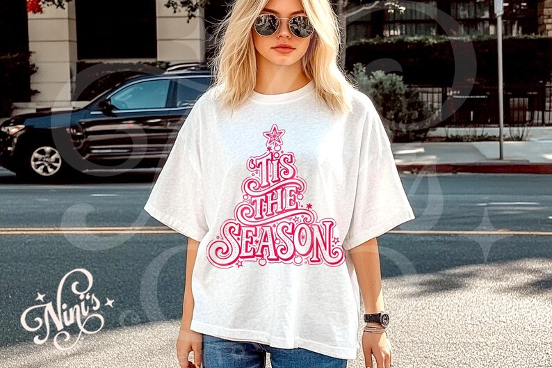 Op de afbeelding: Een wit T-shirt met een roze kerstboomontwerp en de tekst "Tis The Season". Het T-shirt wordt gedragen door een persoon met een zonnebril en een spijkerbroek. Een casual, feestelijk kledingstuk.